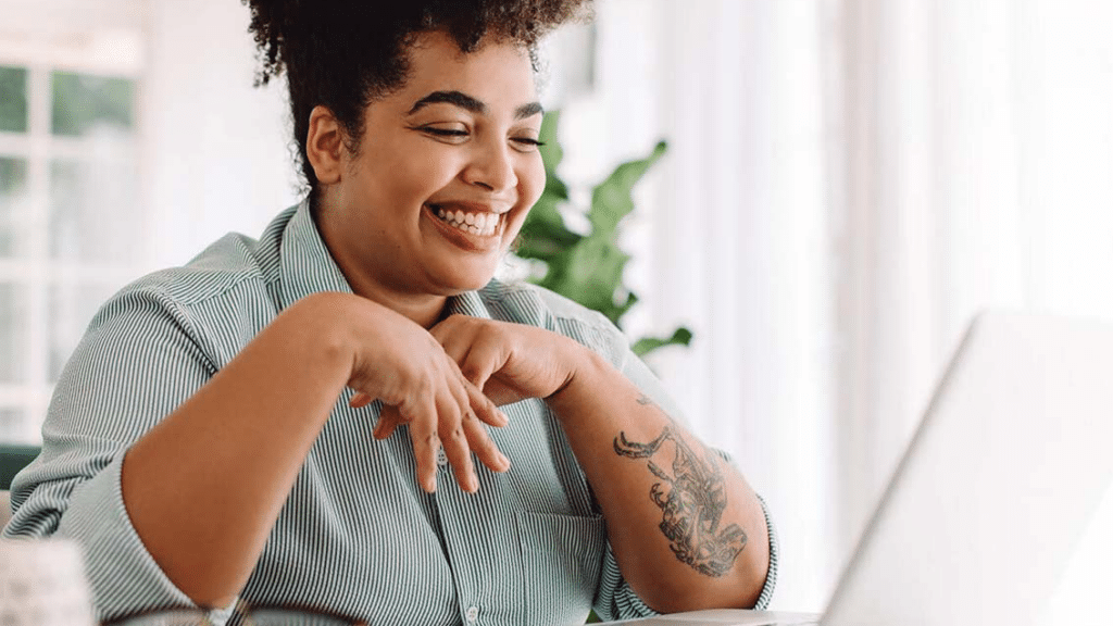 Smiling woman with a tattoo on her arm, sitting at a desk and engaging with a laptop, with natural light and plants in the background