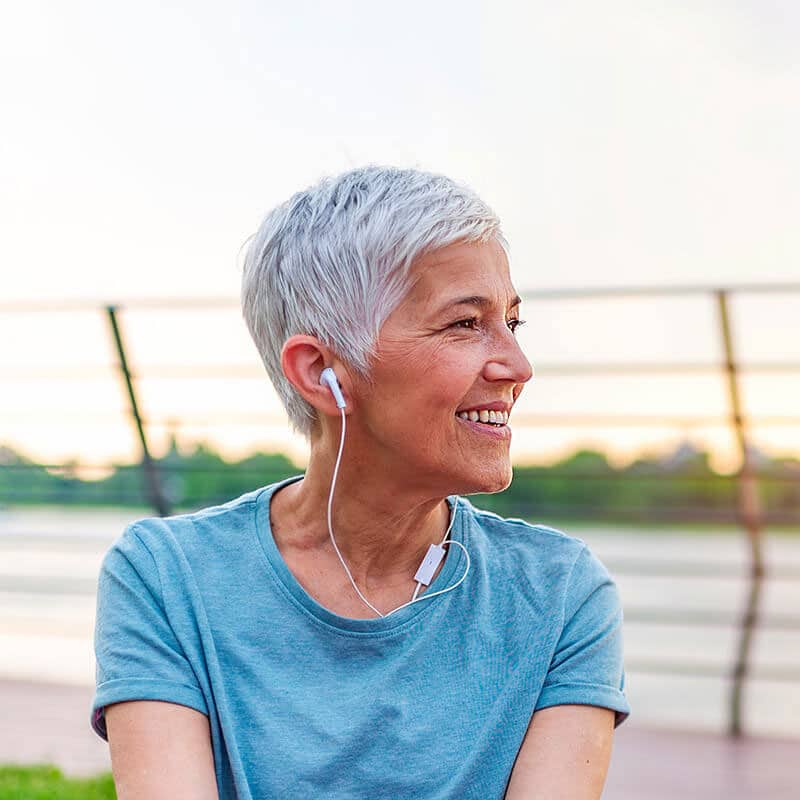 Smiling elderly woman with short gray hair wearing earphones, enjoying the outdoors with a sunset in the background