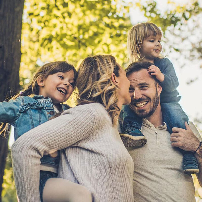 Happy family outdoors, with a father carrying a young child on his shoulders, a mother smiling at him, and another child laughing beside them under a canopy of trees