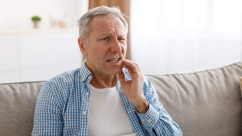 Older man sitting on a sofa, holding his cheek in discomfort from a toothache